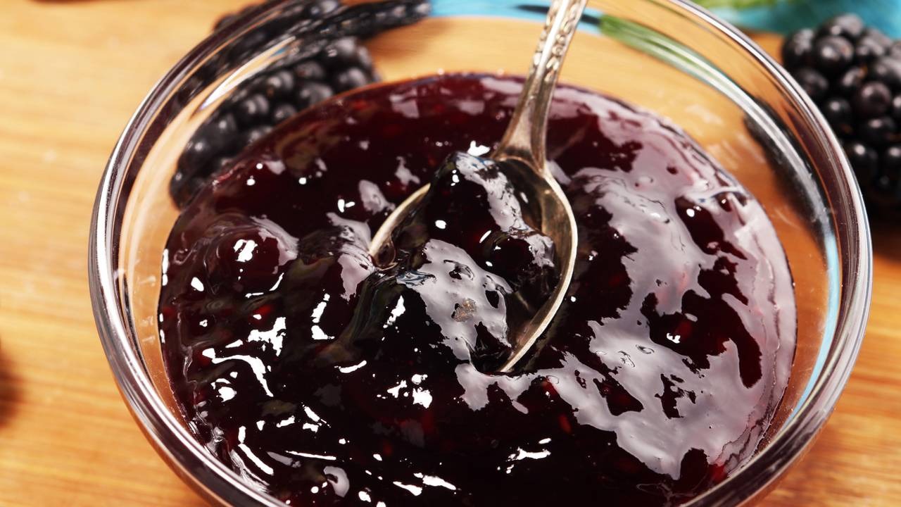 Geleias caseiras de amora são uma das opções para transformar as frutas frescas do quintal em sobremesas sofisticadas e inesquecíveis. Colher de metal em tigela de vidro com geleia escura de amora e frutos inteiros.