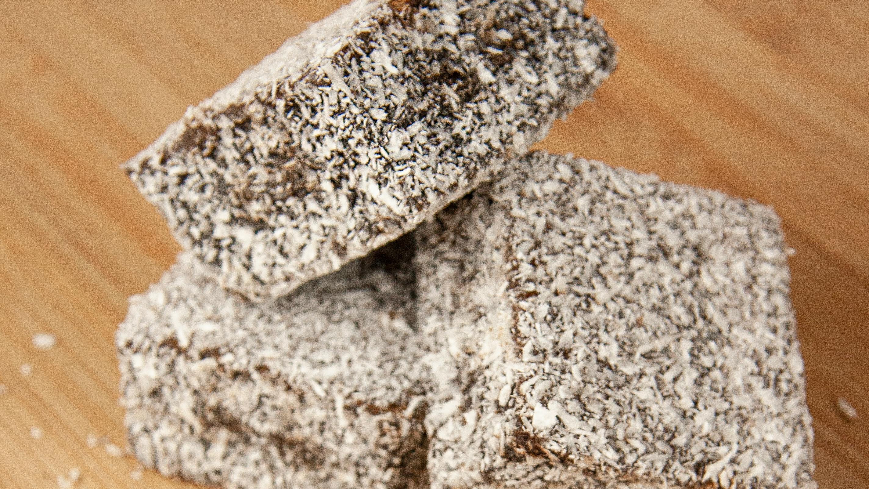 Tr&ecirc;s peda&ccedil;os de bolo retangulares cobertos com coco ralado, sobre uma superf&iacute;cie de madeira clara.