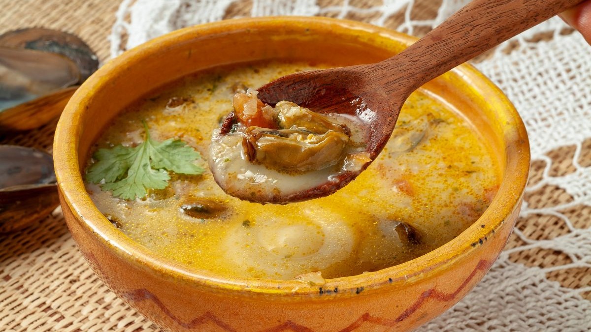 O Caldo de Sururu baiano é uma opção revigorante e saborosa para os dias mais amenos na capital. Sopa cremosa com moluscos e coentro em tigela de cerâmica, com colher de pau.