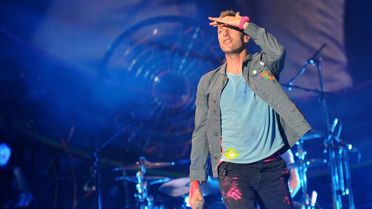 Chris Martin, líder do Coldplay, é o curador do show do intervalo da final da Copa do Mundo de 2026, com participação da banda. Chris Martin no palco, camiseta azul, jaqueta cinza e calça escura com respingos de tinta.