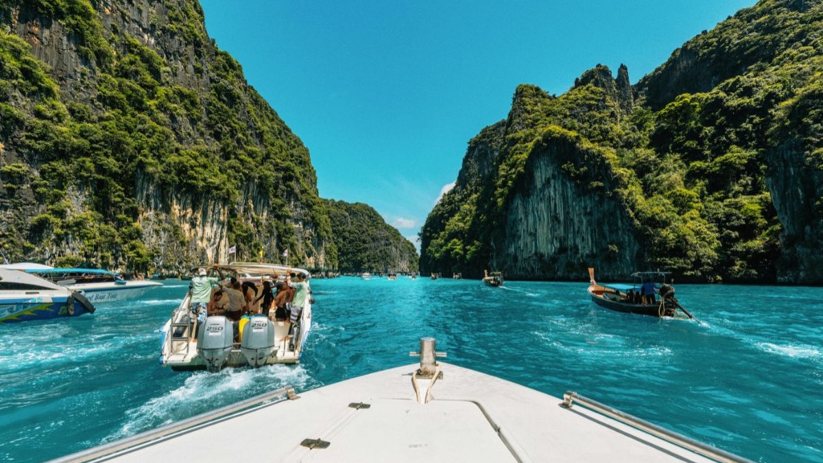 A Tailândia, no Sudeste Asiático, oferece paisagens paradisíacas e custos atraentes para brasileiros com o real valorizado. Vista da proa de um barco em canal de água azul entre falésias verdes com outros barcos.