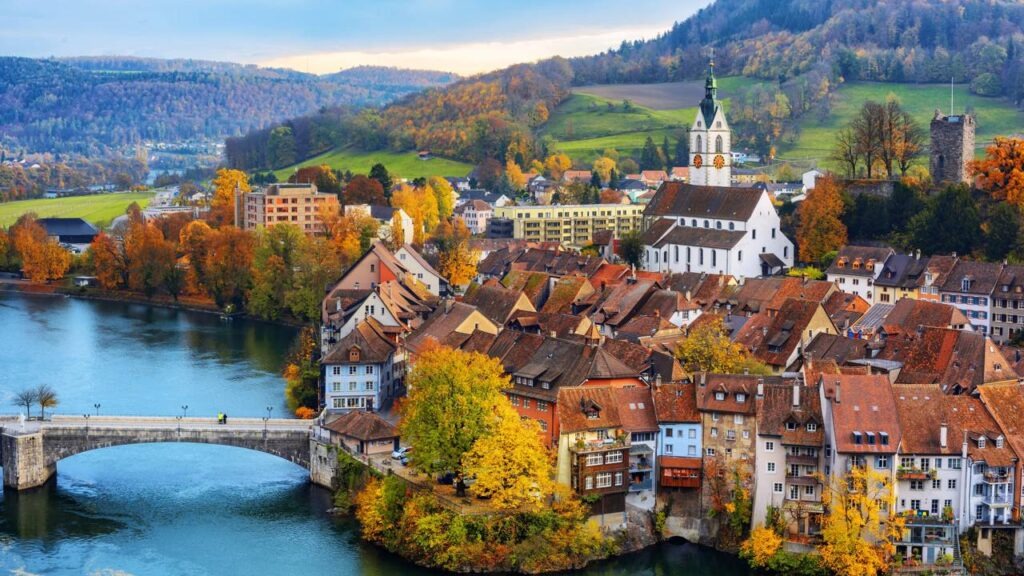 A arquitetura e paisagem suíça ilustram o planejamento urbano e o padrão de vida abordados na reportagem. Vista panorâmica de uma cidade ribeirinha, com casas coloridas, igreja branca, ponte de pedra e florestas em outono.