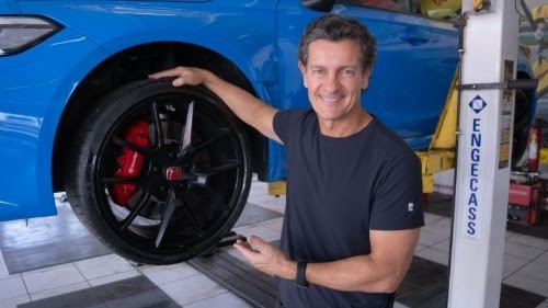 Homem sorridente aponta para pneu de carro azul levantado, com pin&ccedil;a de freio vermelha e elevador ENGECASS