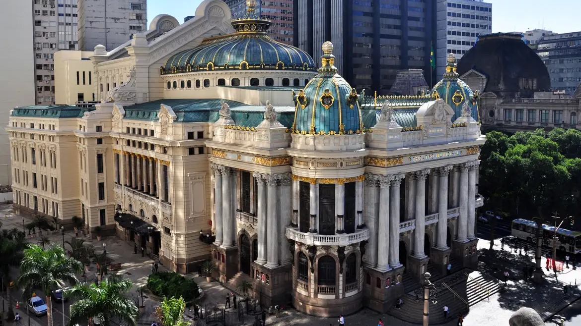 O Theatro Municipal do Rio de Janeiro sedia concerto da Orquestra Sinfônica Jovem, parte da programação cultural. Foto: Riotur/Divulgação Edifício Theatro Municipal com letreiro "THEATRO MUNICIPAL", cúpulas verde-azuladas, detalhes dourados e colunas brancas