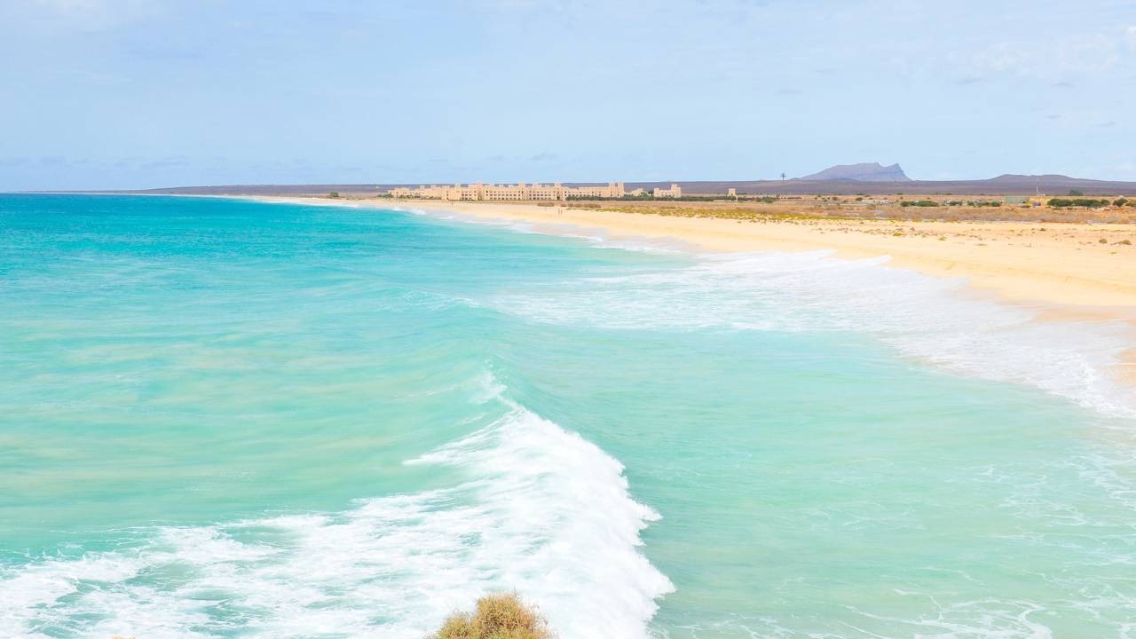 O turismo é o principal motor econômico de Cabo Verde, atraindo visitantes com suas deslumbrantes paisagens costeiras. Mar azul-turquesa com ondas quebrando, praia de areia amarela e complexo de edifícios ao longe.