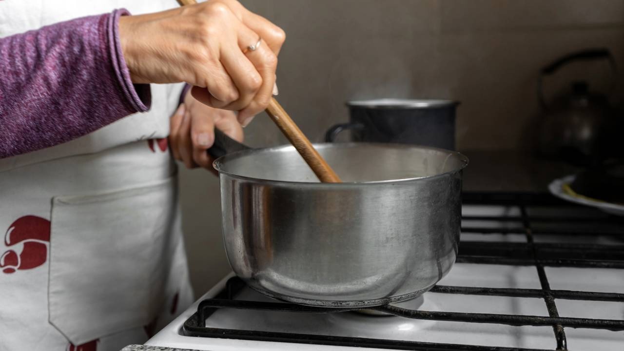 O preparo de caldos quentinhos na cozinha oferece conforto e sabor para enfrentar as baixas temperaturas do inverno. Mãos de pessoa com avental mexendo panela no fogão com colher de pau.
