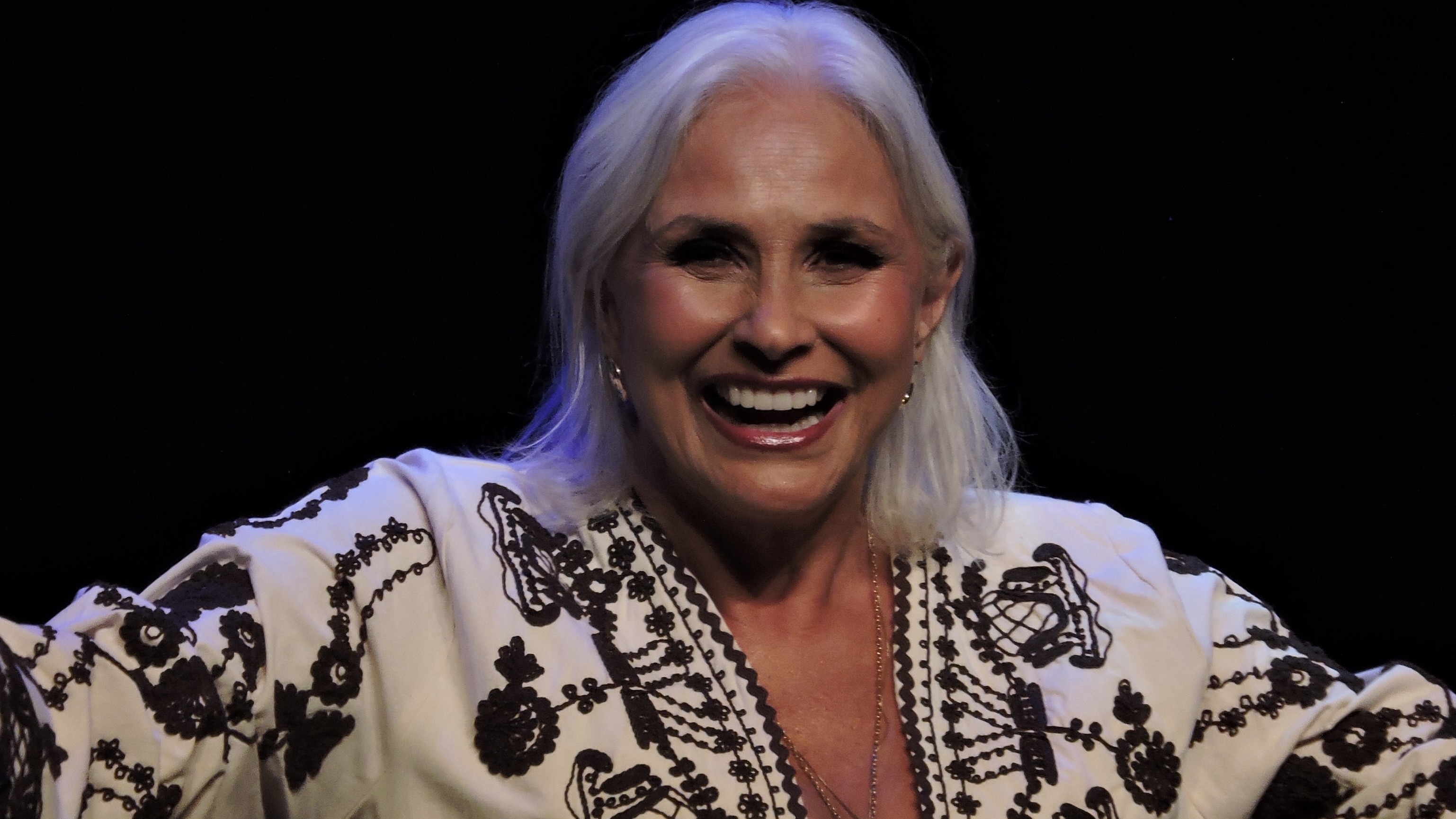 Fafá de Belém celebra 50 anos de carreira com show gratuito nos Arcos da Lapa na Semana de Arte do Rio. Fafá de Belém sorridente com cabelo branco, blusa bordada e colar em palco escuro