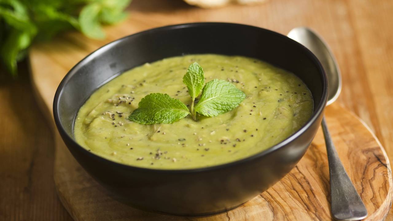 Aprenda a fazer a sopa de ervilha de Bela Gil, uma das opções saudáveis para o inverno. Tigela preta com sopa verde cremosa guarnecida com hortelã e pimenta preta sobre tábua de madeira