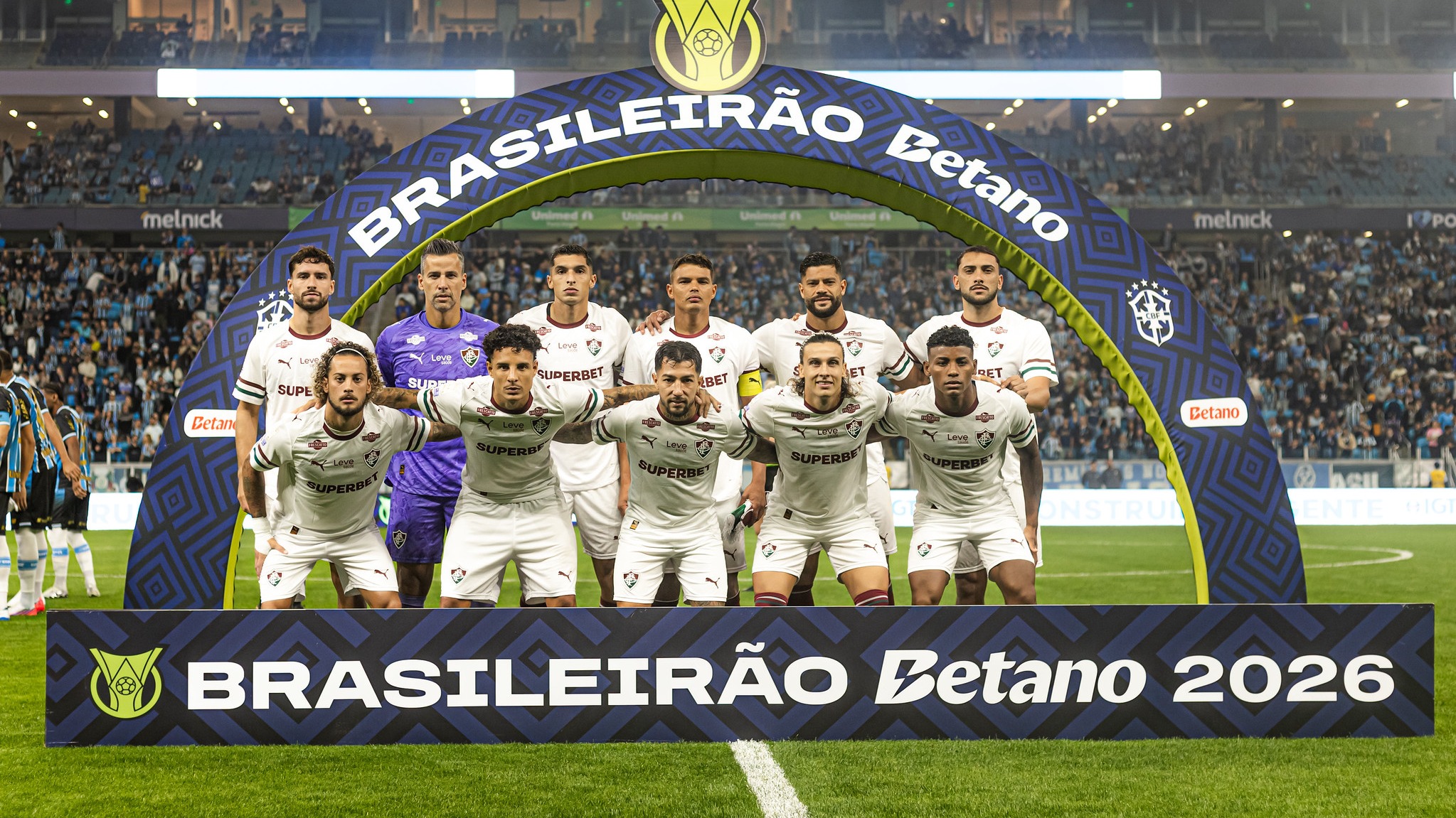 Elenco do Fluminense em formação para os desafios no Brasileirão Betano, Copa do Brasil e Libertadores Time Fluminense posando campo, arco BRASILEIRÃO Betano e sinalização BRASILEIRÃO Betano 2026