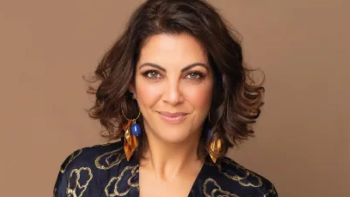 Mulher sorrindo, cabelo castanho-escuro, brincos azuis e dourados, blusa estampada, fundo bege.