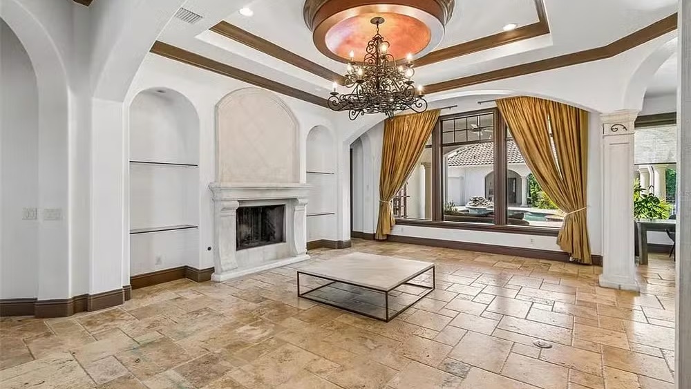 O requinte da sala com piso de travertino e vista para a piscina na mansão de Gugu Liberato, agora à venda - Foto: Divulgação Ampla sala com lareira, piso de travertino, lustre no teto e vista externa para piscina