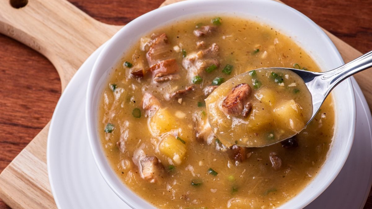 O caldo cremoso, um clássico da culinária mineira, é ideal para aquecer e trazer conforto nas baixas temperaturas do inverno. Caldo cremoso com pedaços de carne, batata e cheiro-verde em tigela branca com colher.
