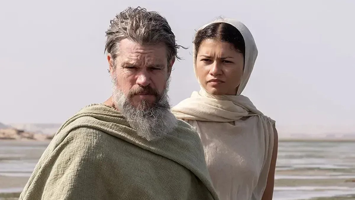 Matt Damon como Odisseu e Zendaya em cena de "A Odisseia", filme épico que recria a jornada do herói grego. Matt Damon, barbudo e de manto, e Zendaya, de véu e túnica, em cenário desértico com água