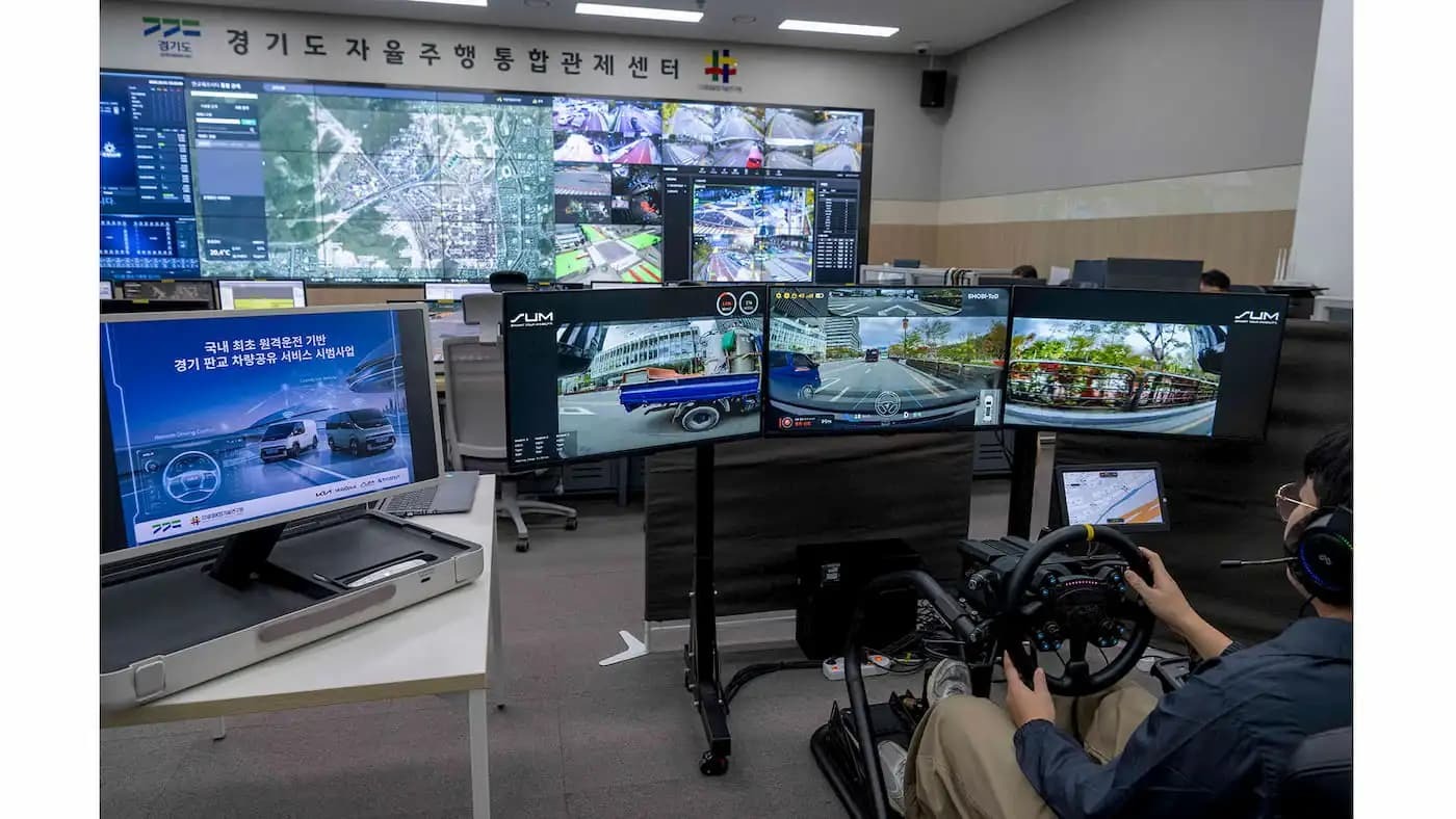 Centro de controle em Gyeonggi, Coreia, gerencia serviço piloto de compartilhamento de vans elétricas da Kia com condução remota Homem em centro de controle com volante, telas exibem ruas e projeto '?? ?? ???? ??? ????'