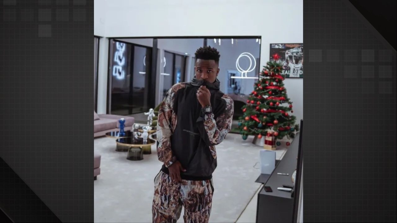 Vini Jr. posa em um dos ambientes de sua mansão em Madrid, imóvel que o jogador exibe frequentemente em suas redes sociais - Foto: Reprodução/Redes Sociais Vini Jr. em moletom estampado, posa na sala com árvore de Natal, relógio e decoração moderna