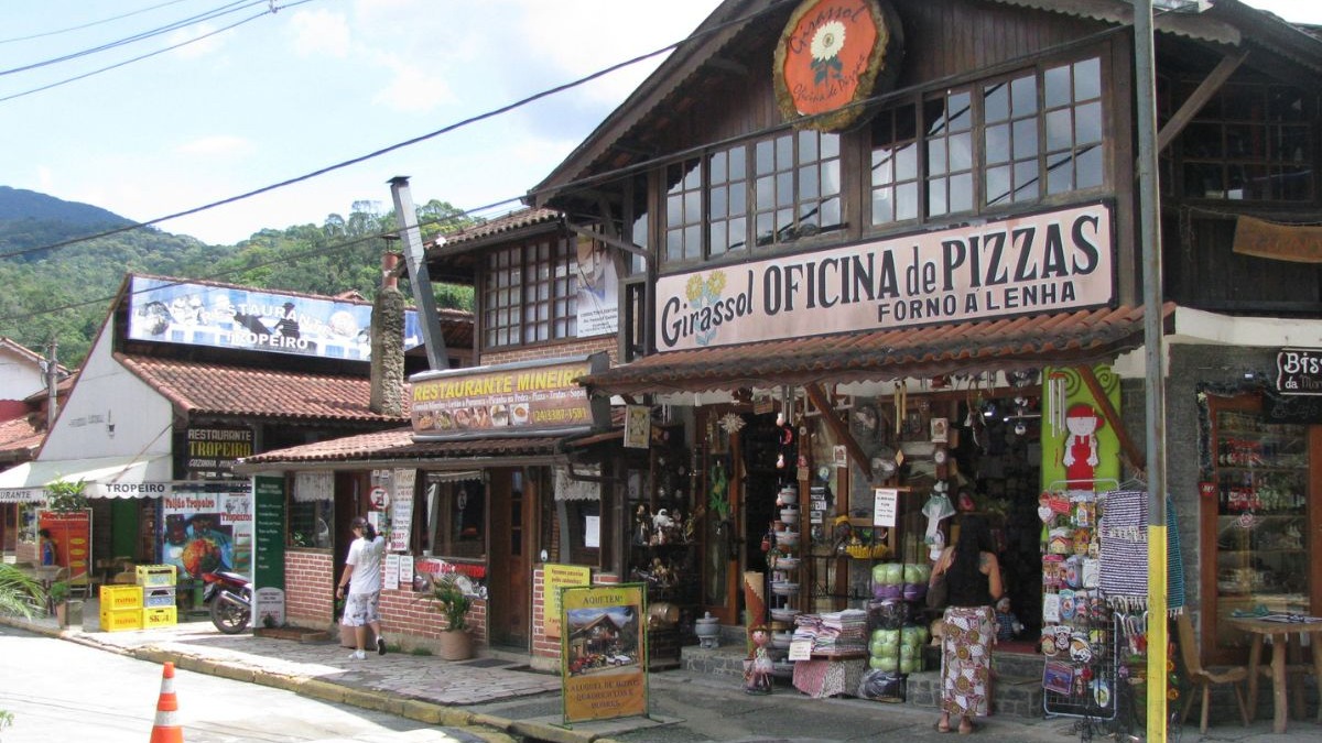 Estabelecimentos e restaurantes mineiros destacam a rica gastronomia das cidades serranas fluminenses. Fachadas de madeira com restaurantes e lojas em rua de cidade serrana com montanhas ao fundo.