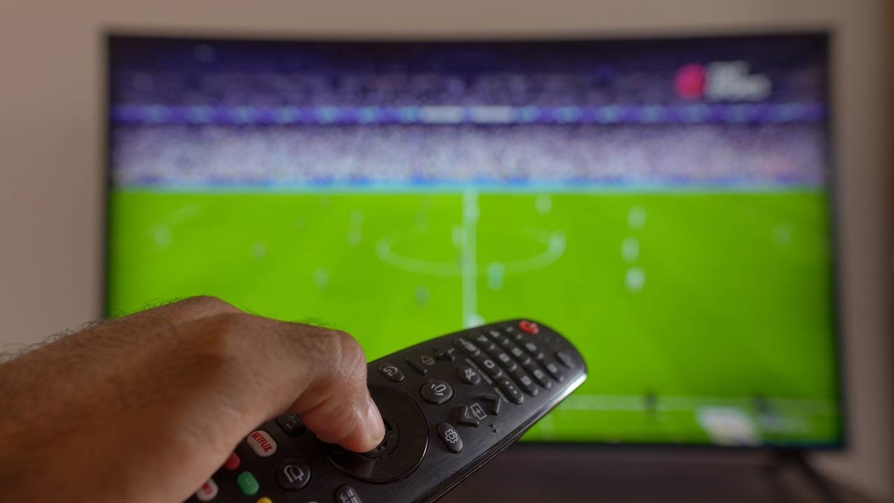 Com petiscos práticos, a Copa do Mundo de 2026 pode ser curtida sem perder um minuto, diretamente do sofá. Mão segura controle remoto de TV com jogo de futebol exibido na tela curva.