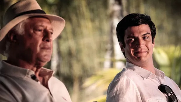 Dois homens em primeiro plano: um mais velho de chapéu e outro mais jovem sorrindo com óculos de sol.