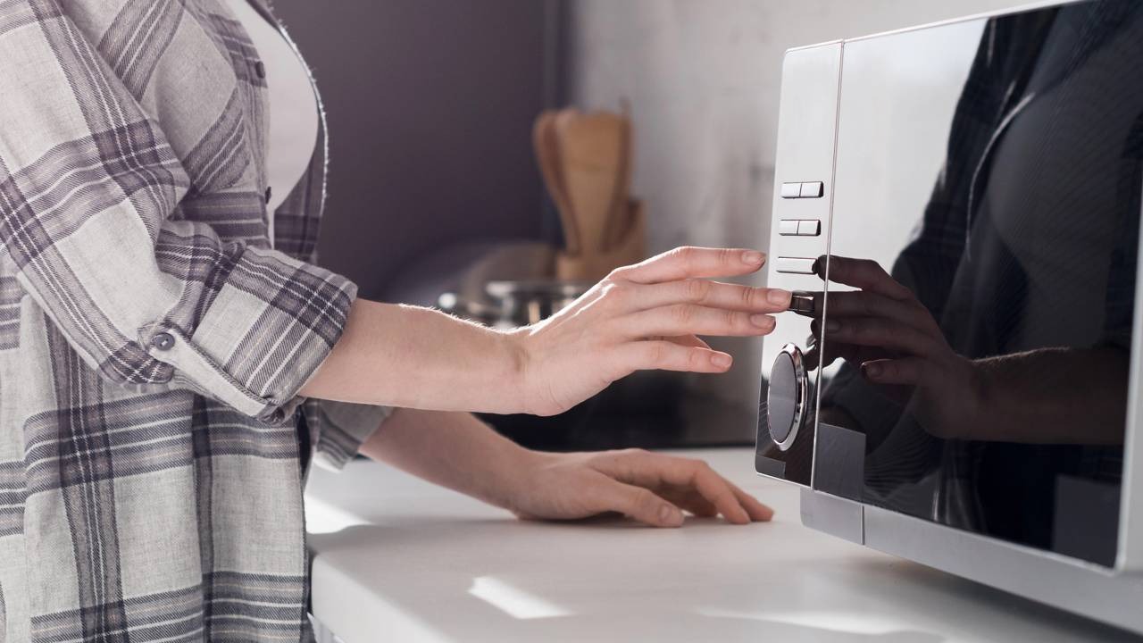 Ajustar o micro-ondas para o teste de potência é um dos passos fundamentais para verificar o bom funcionamento do aparelho. Mãos de pessoa com camisa xadrez ajustam botões de micro-ondas preto e prateado na cozinha