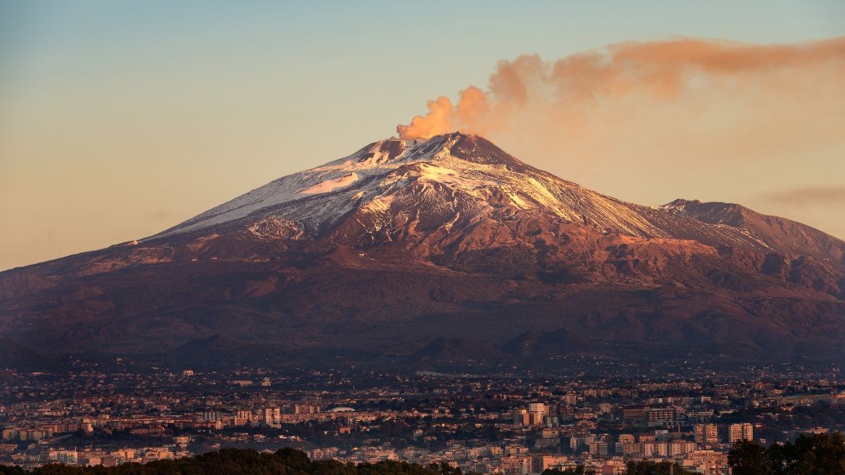O vulcão Etna, na Sicília, exibe sua atividade constante, monitorada para garantir a segurança dos visitantes. Vulcão Etna nevado em erupção com fumaça laranja sobre uma cidade, ao entardecer.