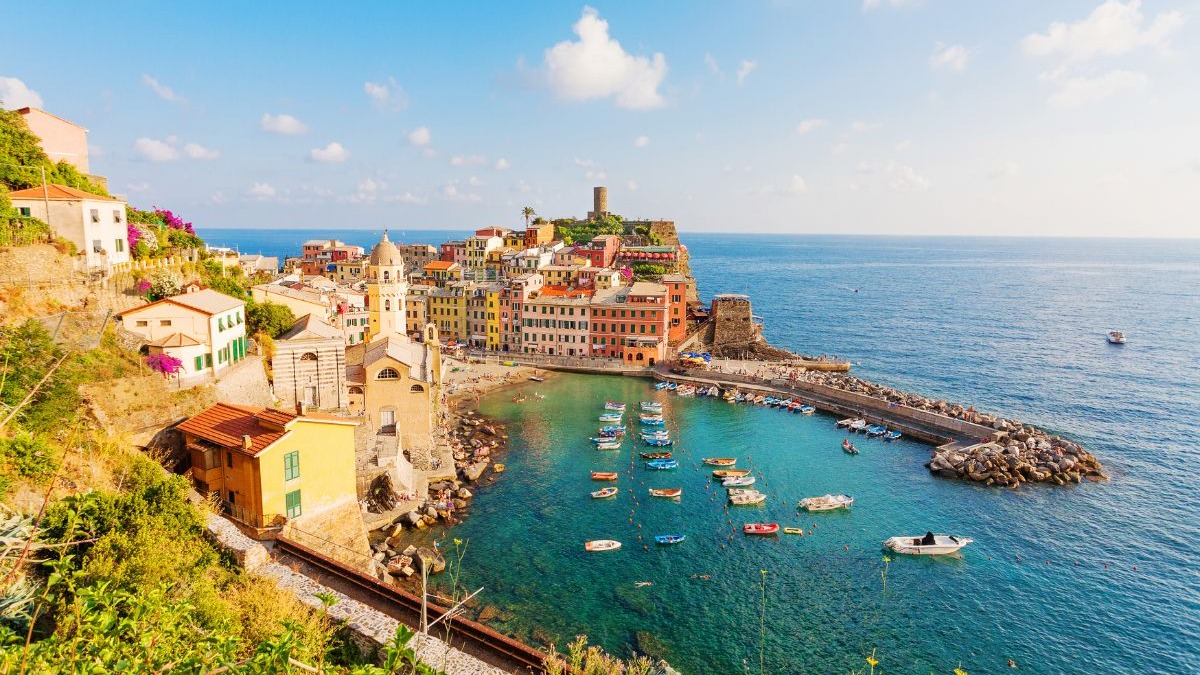 As vibrantes casas coloridas à beira-mar em Cinque Terre ilustram a beleza autêntica dos vilarejos italianos que fogem do óbvio. Vista aérea de vilarejo costeiro com casas coloridas, igreja e barcos em baía azul-turquesa, Itália.