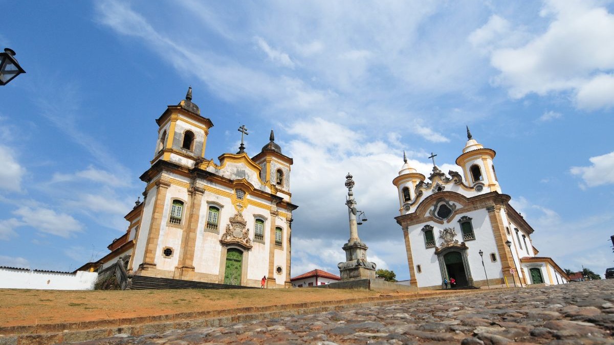 Duas igrejas barrocas coloniais brancas e amarelas e pelourinho em pra&ccedil;a de pedras, c&eacute;u azul.