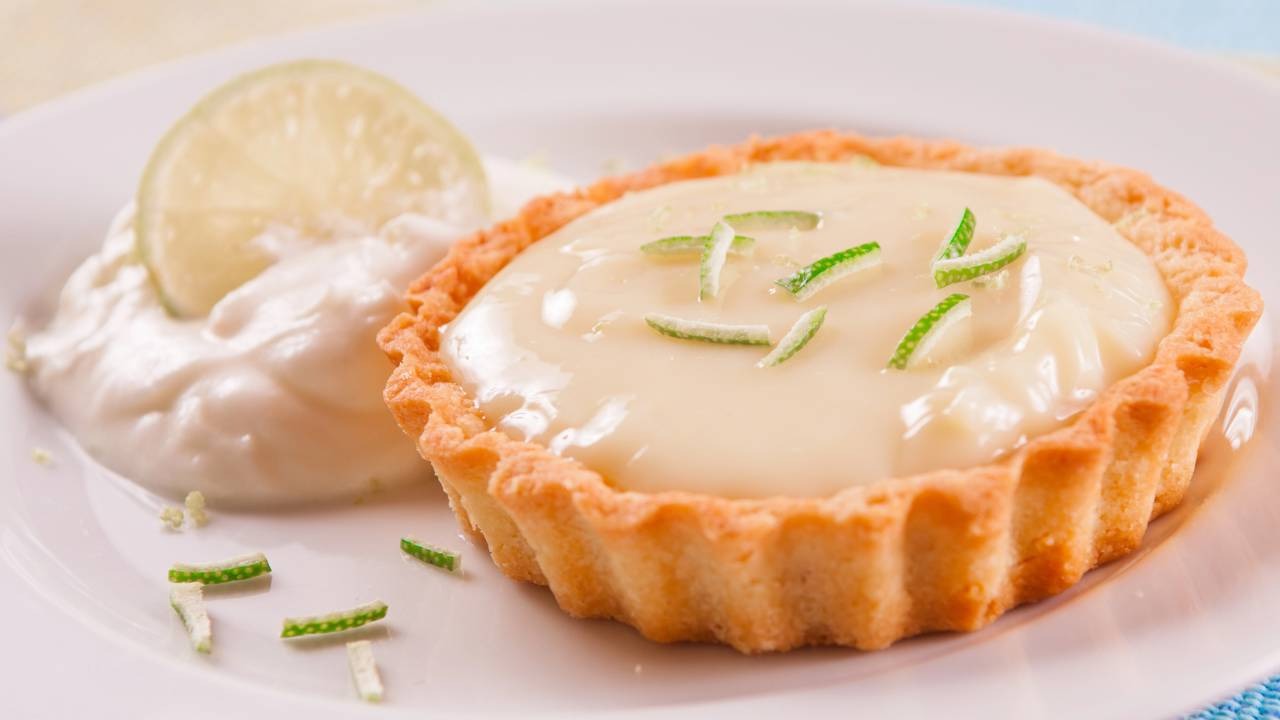 A leveza e o frescor do limão tornam a torta de bolacha uma sobremesa que encanta ao primeiro sabor, perfeita para o dia a dia. Torta individual de limão com raspas verdes, creme branco e uma fatia de limão no prato