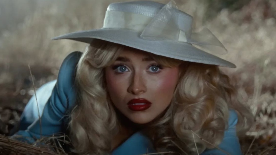 A cantora Sabrina Carpenter em imagem que reflete a estética de seus últimos sucessos, como "Please Please Please" e "Espresso". Foto: Reprodução/YouTube Mulher loira de chapéu branco, olhos azuis e batom vermelho em grama seca.
