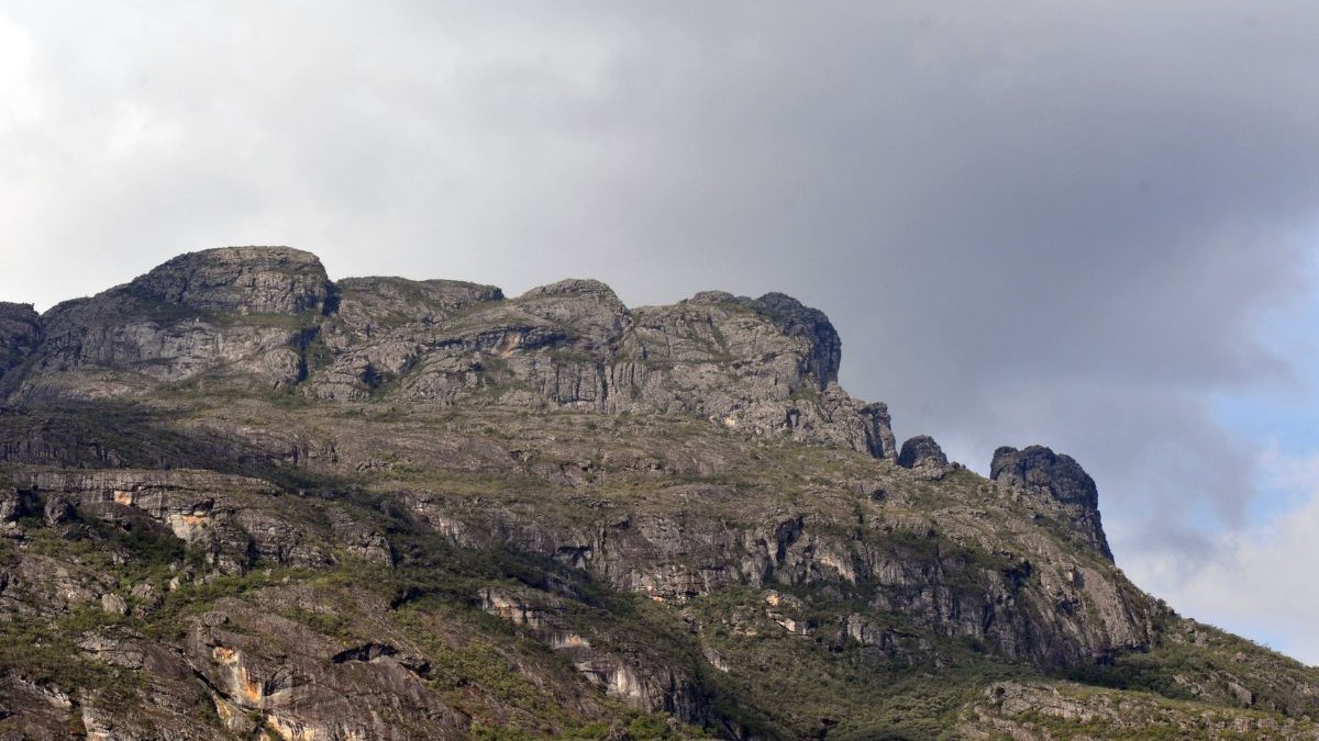 Montanha rochosa cinzenta com cume irregular e vegeta&ccedil;&atilde;o esparsa sob c&eacute;u nublado.