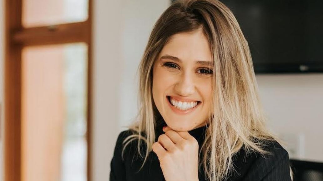 Mulher sorridente de cabelos loiros e blusa preta com a m&atilde;o no queixo
