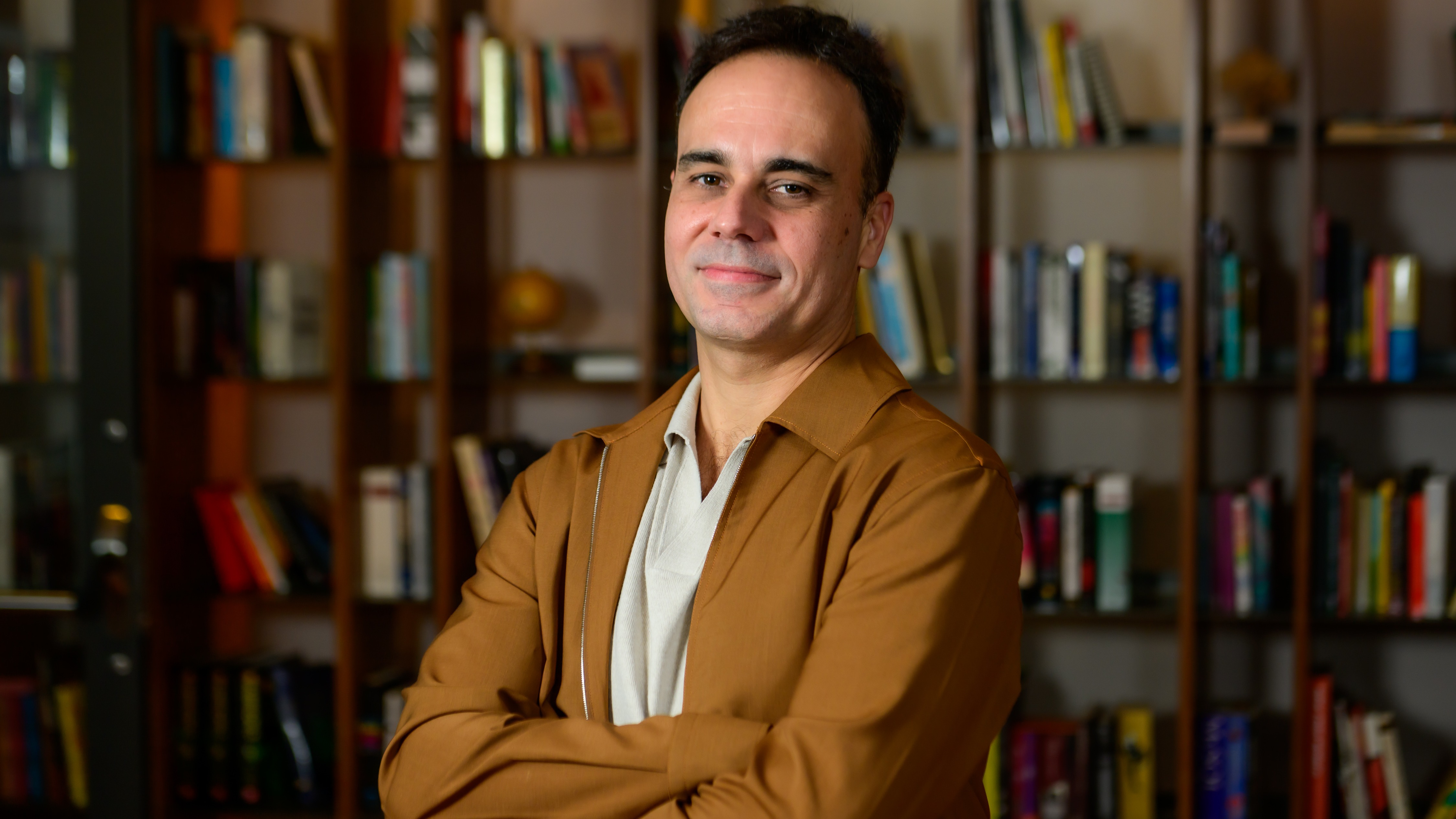 Homem sorridente em jaqueta marrom, bra&ccedil;os cruzados, com estante de livros desfocada ao fundo.