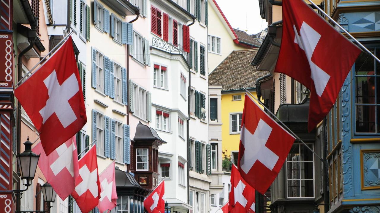 Ruas charmosas da Suíça decoradas com bandeiras nacionais, um cenário comum nos vilarejos de contos de fadas do país. Rua europeia com edifícios coloridos e muitas bandeiras suíças vermelhas com cruz branca.