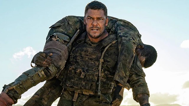 A exigência física dos papéis de ação de Alan Ritchson, evidenciada por sua performance intensa em cena. Homem forte de farda militar carrega outro homem inconsciente sobre os ombros em paisagem desértica
