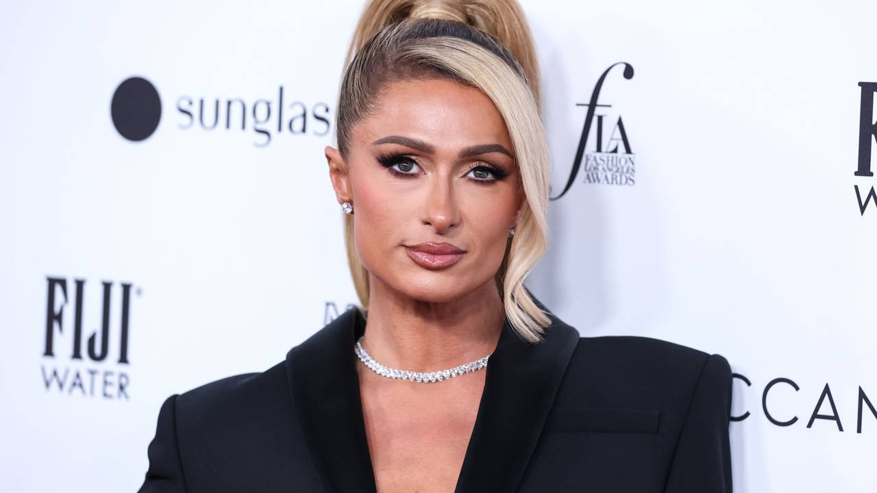 Paris Hilton, que viveu momentos de pânico ao perder um anel de noivado de US$ 2 milhões em uma boate. Paris Hilton com cabelo loiro preso, maquiagem, brincos de diamante e colar choker em fundo branco