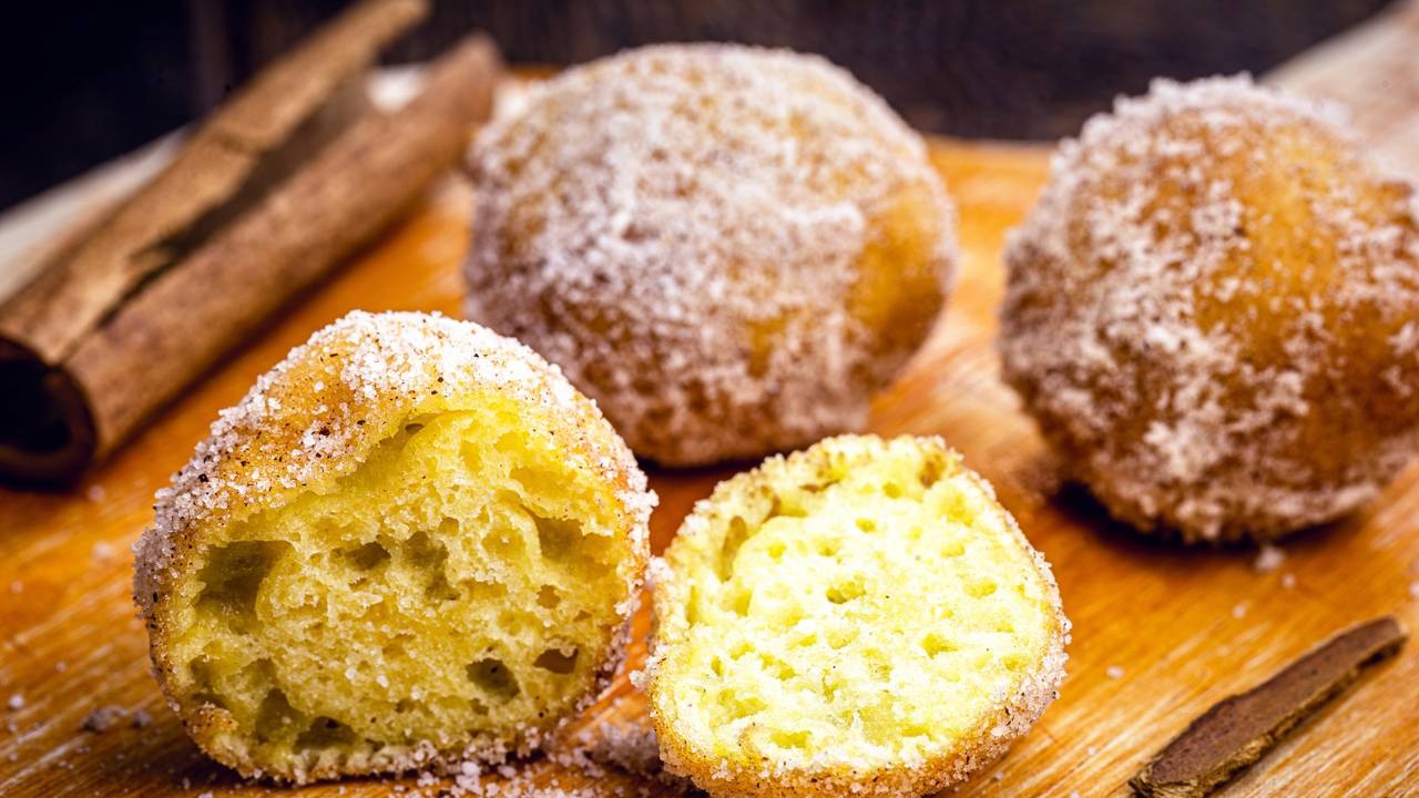 Sabores afetivos do bolinho de chuva com sua massa fofinha e cobertura crocante de açúcar e canela. Bolinhos de chuva polvilhados com açúcar e canela, alguns cortados, em tábua de madeira