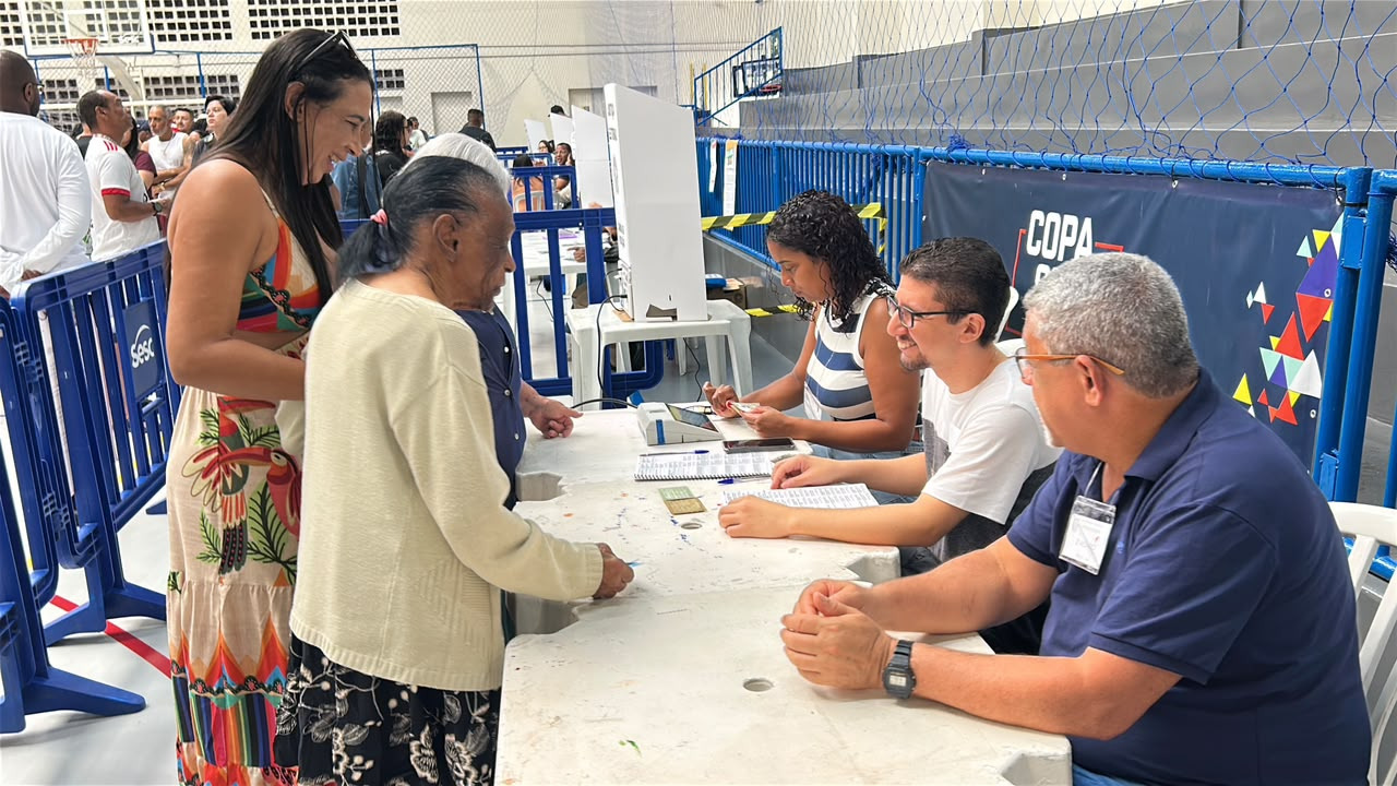 A idosa Maria da Penha, de 102 anos, comparece para votar no Sesc de São João de Meriti, como destaca a matéria - Foto: Alex Machado Idosa vota em seção eleitoral, auxiliada por mulher jovem e mesários em mesa com logo Sesc