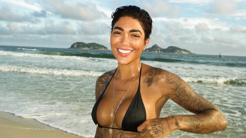 Dhiovanna Barbosa, influencer e irmã de Gabigol, posa na praia, exibindo o corpo e as tatuagens que marcam seu estilo. Foto: Reprodução/Instagram Dhiovanna Barbosa, sorrindo na praia, biquíni preto, tatuagens visíveis e 'Perdon' no pulso