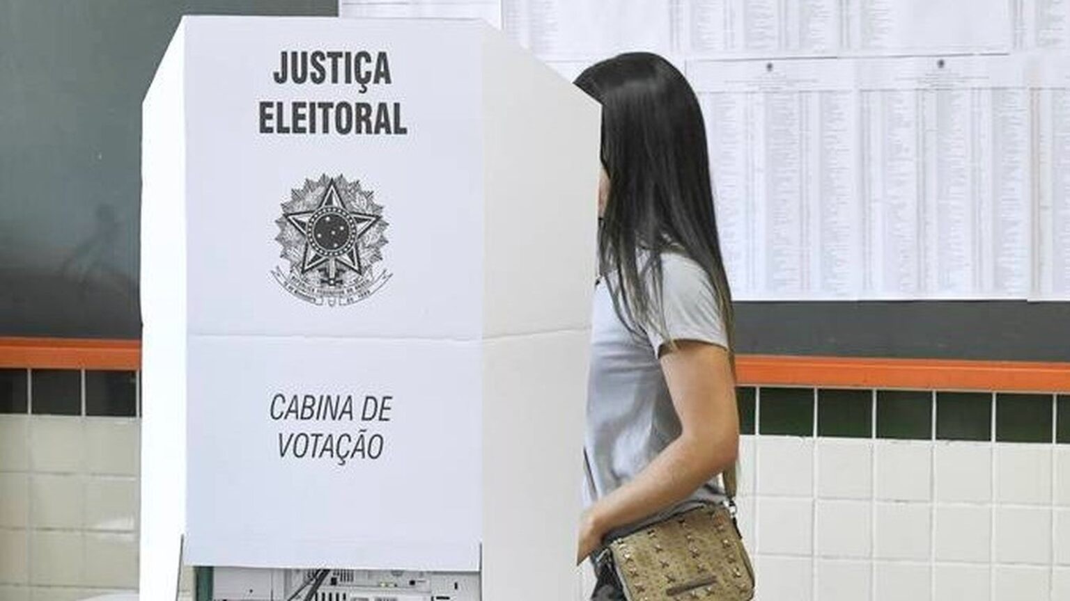 O direito ao voto no segundo turno foi prorrogado em várias seções por falhas no transporte e condições climáticas. Mulher vota em cabina da Justiça Eleitoral; texto diz "JUSTIÇA ELEITORAL", "CABINA DE VOTAÇÃO"