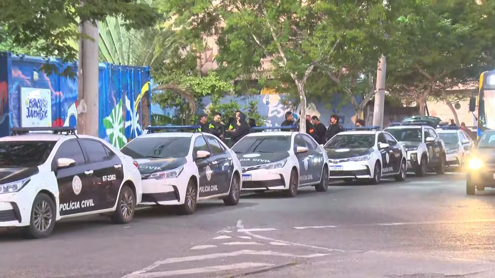 Agentes da Polícia Civil e viaturas participam da Operação Rota Segura Madureira contra a facção TCP no Rio. Viaturas da Polícia Civil e agentes em rua, com contêiner e árvores ao fundo