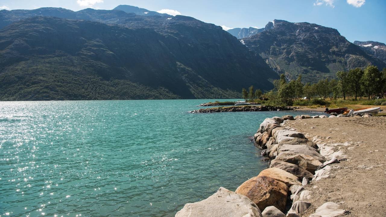 A vasta natureza norueguesa, com lagos e montanhas imponentes, reflete a cultura do país e o direito de acesso universal à terra. Lago azul-turquesa, montanhas arborizadas ao fundo, costa rochosa e praia com pequenos barcos.
