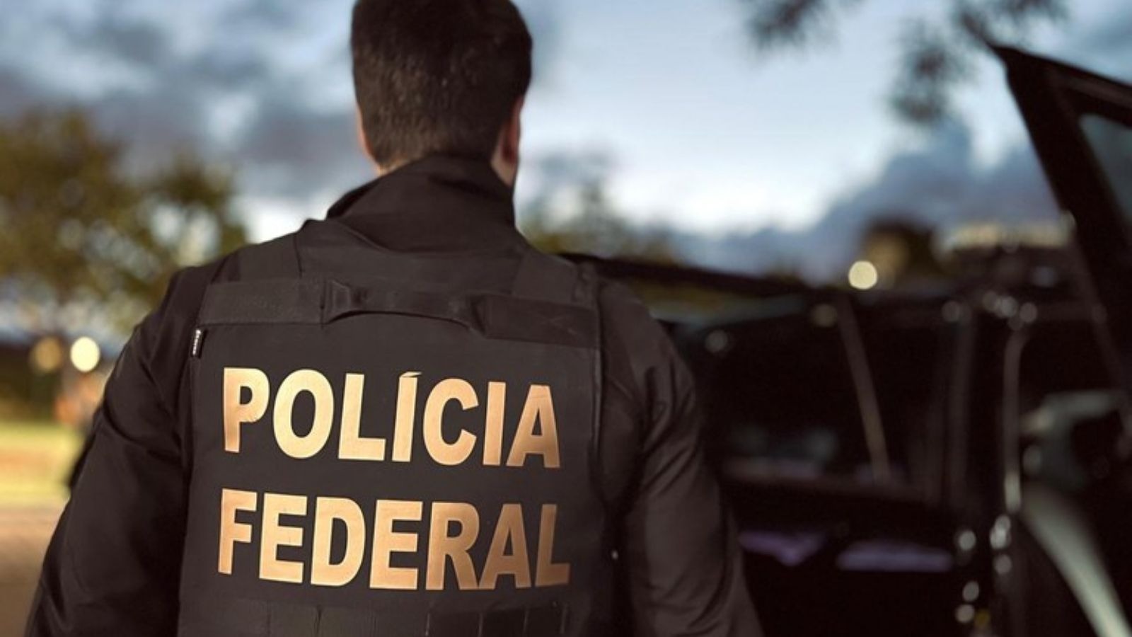 O uniforme da Polícia Federal, corporação que recomendou a demissão de Eduardo Bolsonaro por abandono de cargo. Pessoa de costas em colete preto com "POLÍCIA FEDERAL" em amarelo. Fundo desfocado com carro
