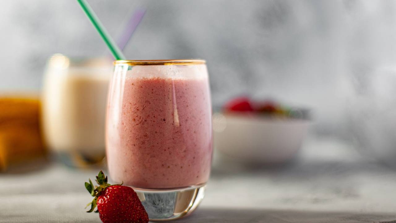 O smoothie de frutas vermelhas com iogurte pode ser uma alternativa prática e saborosa para o café da manhã de sábado. Smoothie rosa em copo com canudo verde e morango fresco no primeiro plano