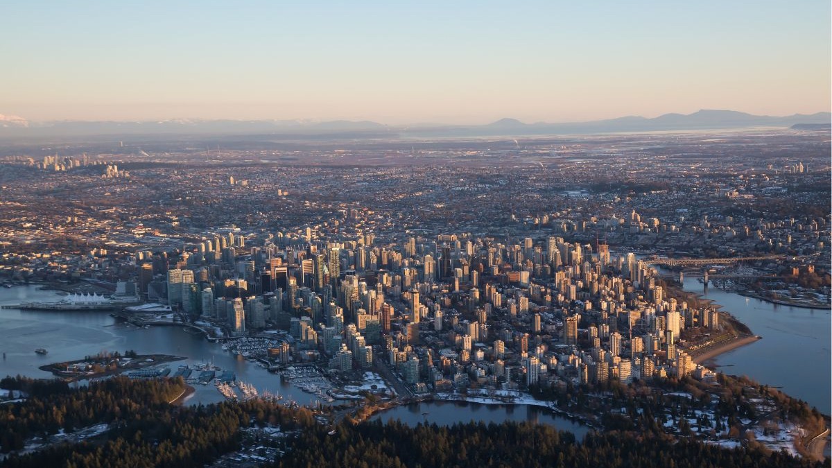Vista a&eacute;rea panor&acirc;mica de Vancouver ao entardecer, com arranha-c&eacute;us, &aacute;gua, floresta e montanhas distantes.