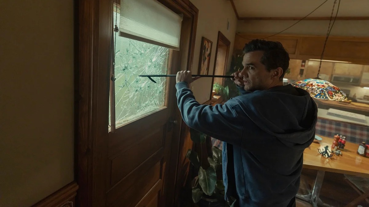 Wagner Moura, elogiado por sua atuação em ‘A Última Casa’, em cena que reflete o suspense do filme da Netflix. Foto: Chris Baker/Netflix Homem de moletom azul segura pé de cabra na frente de porta com vidro quebrado