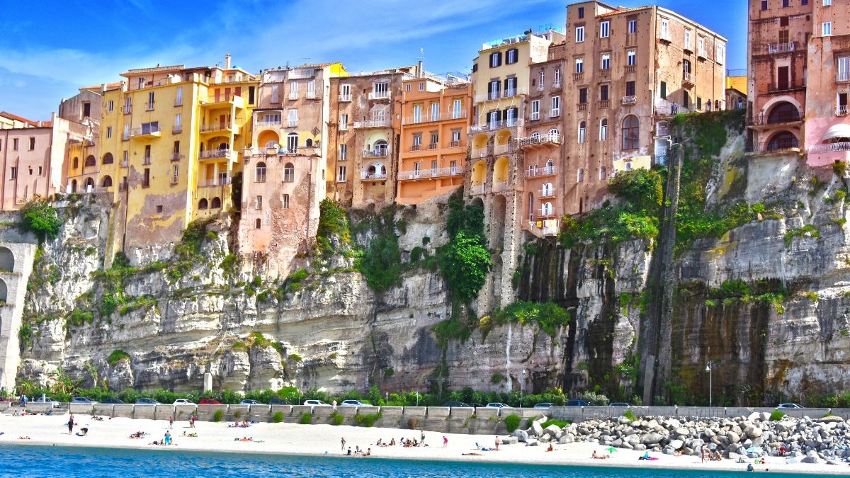 Tropea, na Calábria, destaca-se por suas casas coloridas sobre falésias e praias de águas cristalinas, ideal para uma nova vida na Itália. Casas coloridas em falésias íngremes acima de uma praia de areia e mar azul na Itália.