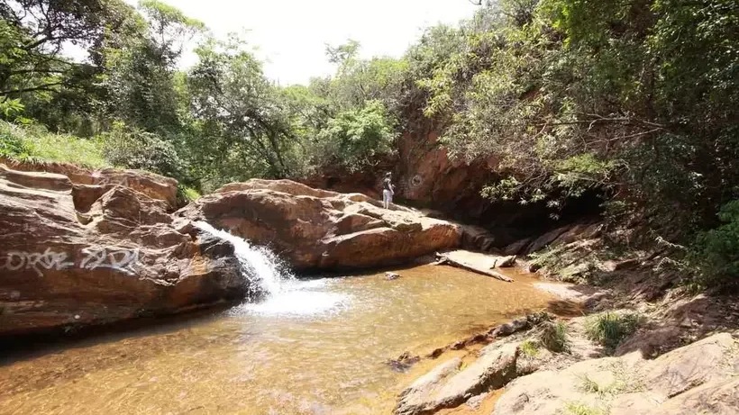 Cachoeira em rochas com grafite, água avermelhada, vegetação verde e pessoa em pé.