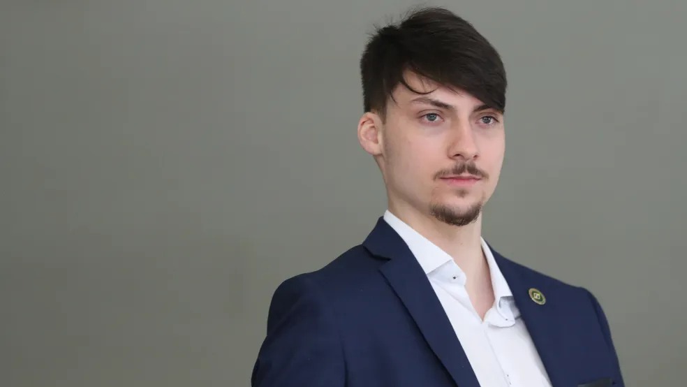 Homem jovem com cavanhaque e bigode, terno azul escuro, camisa branca, olhando para a esquerda