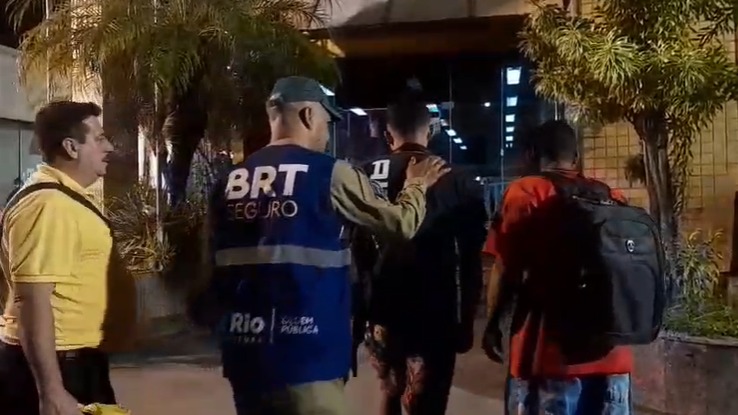 Agentes do programa BRT Seguro atuam na prisão de indivíduo por vandalismo em estação da Zona Oeste - Foto: Reprodução Agente do BRT Seguro em colete azul escolta homem enquanto outros observam em estação noturna