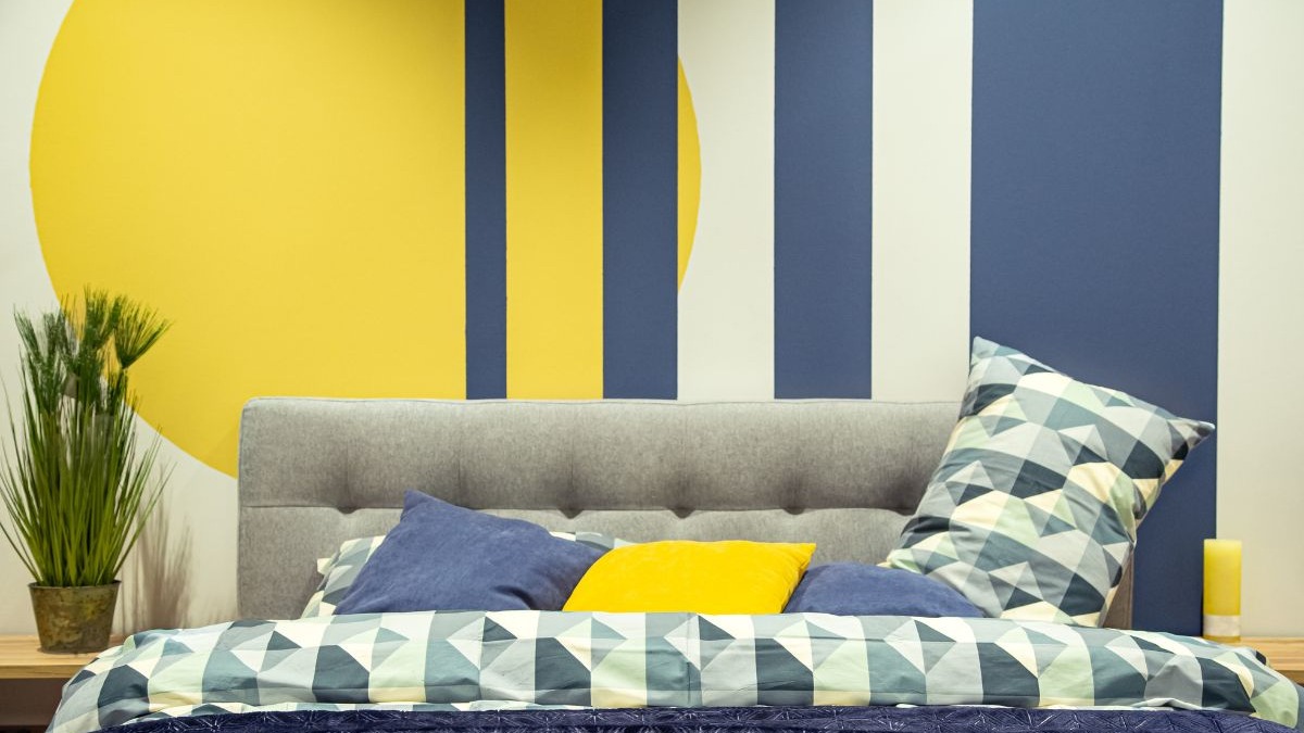 Pinturas criativas na parede, como círculos e listras, substituem a cabeceira física, alinhando-se às tendências de design de 2026. Cama cinza com parede pintada em círculo amarelo e listras azuis e brancas, com planta.