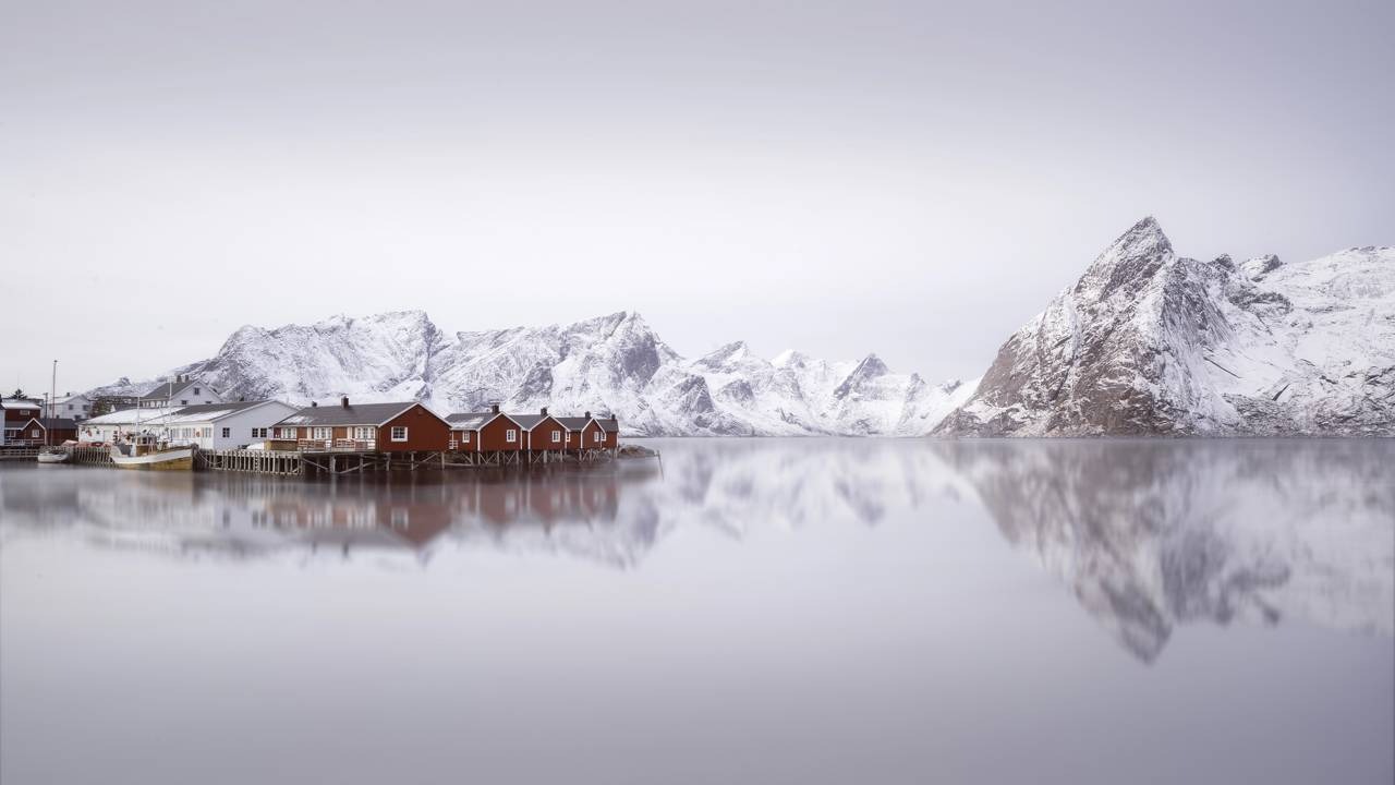 A beleza natural da Noruega, rica em paisagens deslumbrantes, como esta, é palco de fenômenos únicos como o sol da meia-noite. Casas vermelhas e brancas, montanhas nevadas, reflexo na água calma da Noruega.